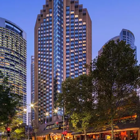 Four Seasons Hotel Sydney Exterior photo The photo shows a tall modern building with a glass façade, likely an office or residential space, illuminated in the evening. In the foreground, there are trees and streetlights, contributing to a lively urban atmosphere. Surrounding the main buildi