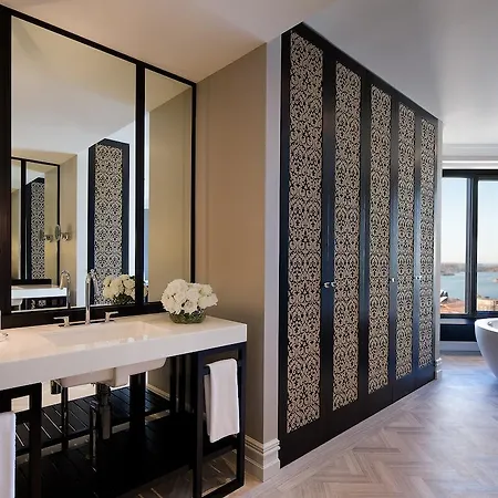 Four Seasons Hotel Sydney Room photo The photo shows a modern and stylish bathroom. It features a double vanity with a large mirror above it, surrounded by decorative panels. The vanity has a sleek white countertop, and there are two sinks. On the countertop, there are white flowers in 