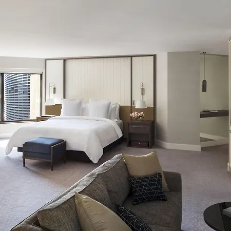 Four Seasons Hotel Sydney Exterior photo The photo shows a modern hotel room featuring a spacious layout. In the center, there's a large bed with white linens, flanked by nightstands with lamps. To one side, a comfortable sofa is positioned, complemented by a small coffee table. There is a 