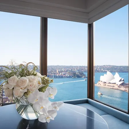 Four Seasons Hotel Sydney Exterior photo The photo shows an elegant interior space with large windows that provide a stunning view. In the foreground, there is a stylish table topped with a beautiful floral arrangement consisting of white roses and orchids in a clear vase. Through the windo