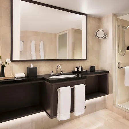 Four Seasons Hotel Sydney Exterior photo The image shows a modern bathroom featuring neutral-colored tiles. There is a large mirror above a dark countertops with a sink, which has various toiletries arranged on it. A small vase with a flower adds a decorative touch. Two white towels hang on