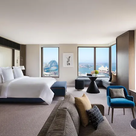 Four Seasons Hotel Sydney Exterior photo The photo shows a spacious hotel room with modern decor. It features a large bed with white linens, positioned against a wall with a contemporary headboard. There are large windows offering a view of an outdoor scene, possibly featuring a landmark li
