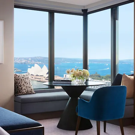 Four Seasons Hotel Sydney Exterior photo The photo shows a stylish hotel room with a large window offering a view of the water and the Sydney Opera House in the distance. There is a small, modern dining table with a vase of flowers on it, situated near the window. A comfortable blue chair i