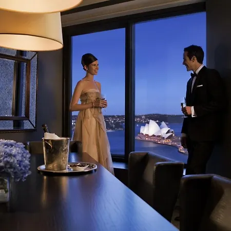 Four Seasons Hotel Sydney Exterior photo The photo depicts a stylish evening scene where a woman in an elegant dress and a man in a formal suit are engaged in conversation near a large window. They are in a modern interior space, with a table that has a bottle of champagne and a glass. The 