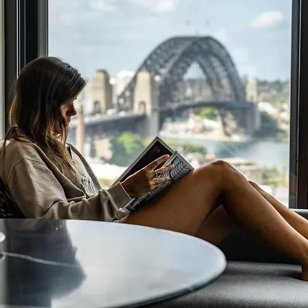 Four Seasons Hotel Sydney Exterior photo The photo depicts a woman sitting comfortably by a window, reading a book. She is wearing a casual oversized sweatshirt and is relaxed, with her legs crossed. The background features a view of a bridge, likely the Sydney Harbour Bridge, with a citysc