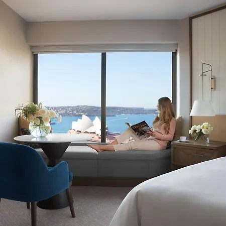 Four Seasons Hotel Sydney Exterior photo The photo shows a stylish hotel room with a large window offering a view of a scenic waterfront, likely featuring notable architecture, such as the Sydney Opera House. A woman is seated on a window bench, reading a book or magazine. The room is elega
