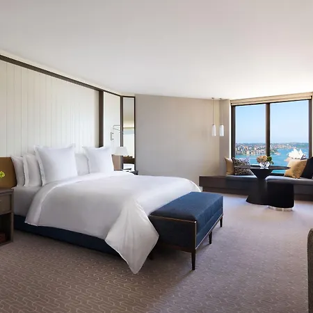 Four Seasons Hotel Sydney Exterior photo The photo shows a modern hotel room that features a spacious layout. In the center, there is a large bed with white linens, complemented by a blue upholstered bench at the foot of the bed. The walls have a light and neutral color scheme, creating a c