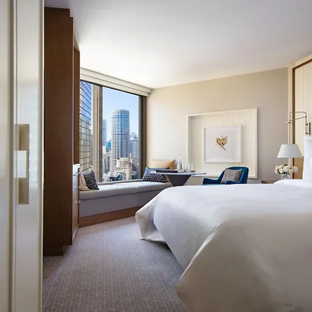 Four Seasons Hotel Sydney Exterior photo The photo shows a modern hotel room with a large bed, featuring a crisp white duvet and pillows. There is a stylish seating area with a cozy window nook and comfortable chairs, allowing for a view of the city skyline outside. The room has contemporar