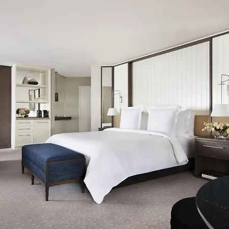 Four Seasons Hotel Sydney Exterior photo The photo depicts a modern and elegantly designed hotel room. It features a spacious layout with a large bed covered in white bedding, flanked by bedside tables and lamps. There is a stylish blue ottoman at the foot of the bed, adding a touch of colo