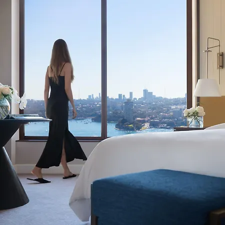Four Seasons Hotel Sydney Exterior photo The photo depicts a stylish hotel room with a woman standing near a large window. She is wearing a black dress and appears to be admiring the view of a cityscape, which includes water and buildings in the background. The room features modern decor, w