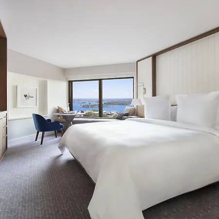 Four Seasons Hotel Sydney Exterior photo The photo shows a modern hotel room designed with a clean and elegant aesthetic. The room features a large bed with white linens and multiple pillows. There is a blue chair positioned near a window that offers a view of the outdoors, likely showcasin