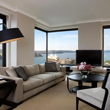 Four Seasons Hotel Sydney Exterior photo The photo depicts a modern and elegantly designed living room with large windows offering a panoramic view of a waterfront scene. The room features a light-colored sofa with decorative cushions, a round coffee table in the center, and a stylish floor
