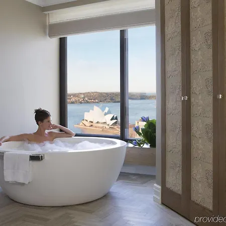 Four Seasons Hotel Sydney Exterior photo The image shows a modern bathroom featuring a large white freestanding bathtub. A person is sitting in the tub, enjoying a bath with bubbles. In the background, there's a large window that offers a view of a body of water and a landmark. The walls ar