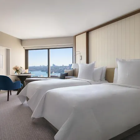 Four Seasons Hotel Sydney Exterior photo The photo shows a contemporary hotel room featuring two neatly made beds with white linens. There is a cozy blue chair positioned near a table, and a vase with flowers on the table adds a touch of decor. The large window offers a view of the outside,